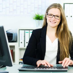 Junge Frau mit langen Haaren und Brille sitzt vor dem Computer am Schreibtisch